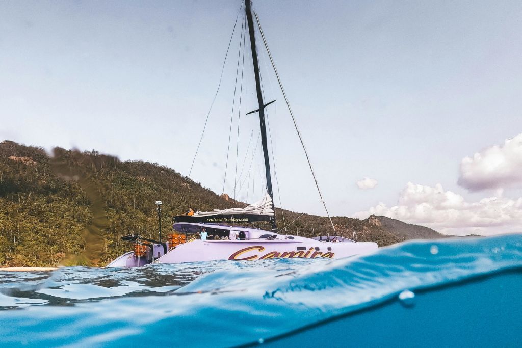 "카미라" 라는 이름의 요트는 숲이 우거진 해안선 근처의 잔잔한 물에 정박해 있으며, 돛대는 부분적으로 흐린 하늘로 뻗어 있습니다.