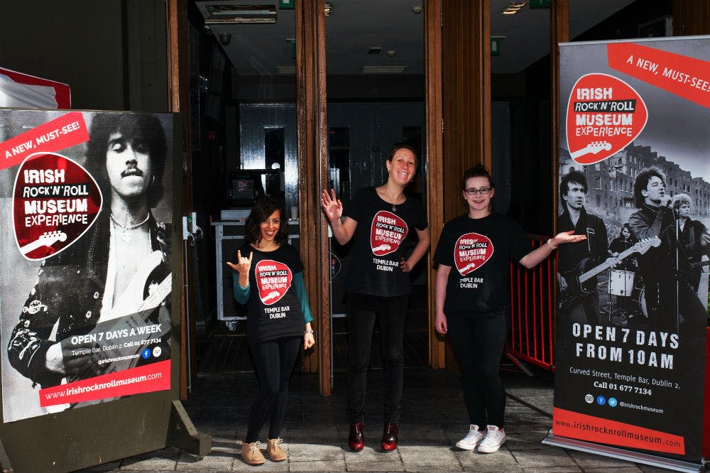 아일랜드 로큰롤 박물관 체험 표지판 옆에 어울리는 티셔츠를 입은 세 사람이 서 있습니다.