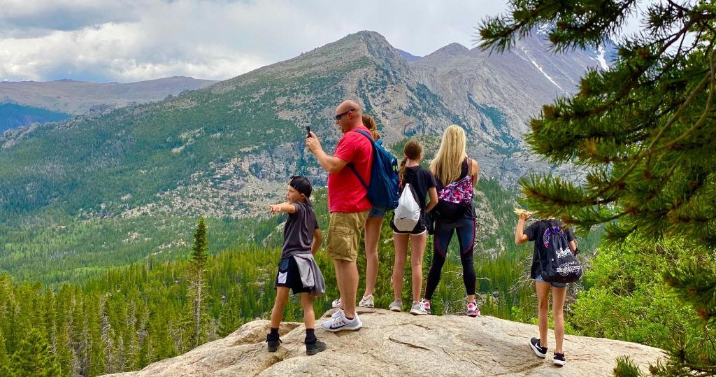 어른과 어린이를 포함한 한 무리의 사람들이 바위 위에 서서 산이 우거진 숲 풍경을 내려다보고 있습니다.