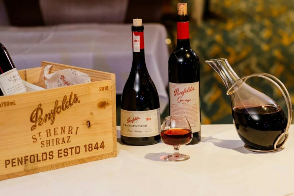 흰색 식탁보 위에 펜폴드 와인 2병, 나무 상자, 와인 한 잔, 레드 와인이 담긴 디켄터가 놓여 있습니다.