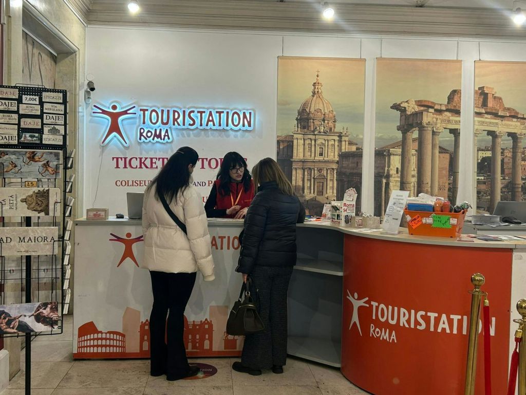'투어리스테이션 로마 (Touristation Roma)' 라고 적힌 관광 안내 카운터에 3명이 모였으며, 홍보 디스플레이와 로마식 건축 장식이 갖춰져 있습니다.