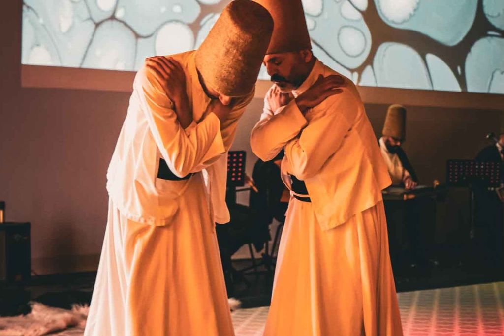 전통 복장을 한 두 사람이 따뜻한 조명으로 꾸며진 방에서 팔짱을 끼고 의식적인 춤을 추고 있습니다.