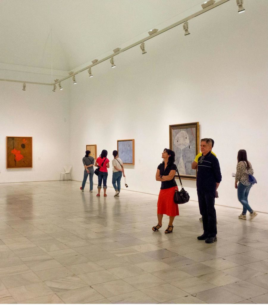 흰색 벽과 타일 바닥이 있는 넓고 조명이 밝은 미술관에서 그림을 감상하는 사람들.