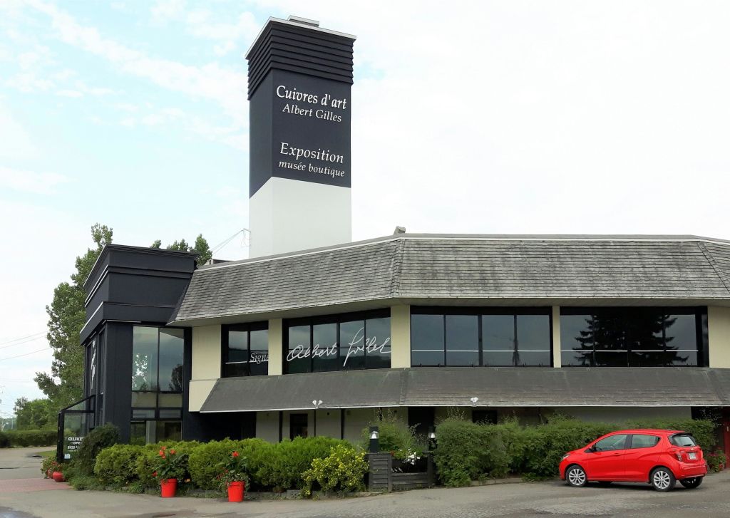 박물관 부티크가 있는 "Cuivres d 'art Albert Gilles" 라고 쓰인 높은 표지판이 있는 건물입니다. 앞에 덤불과 빨간색 자동차가 있습니다.