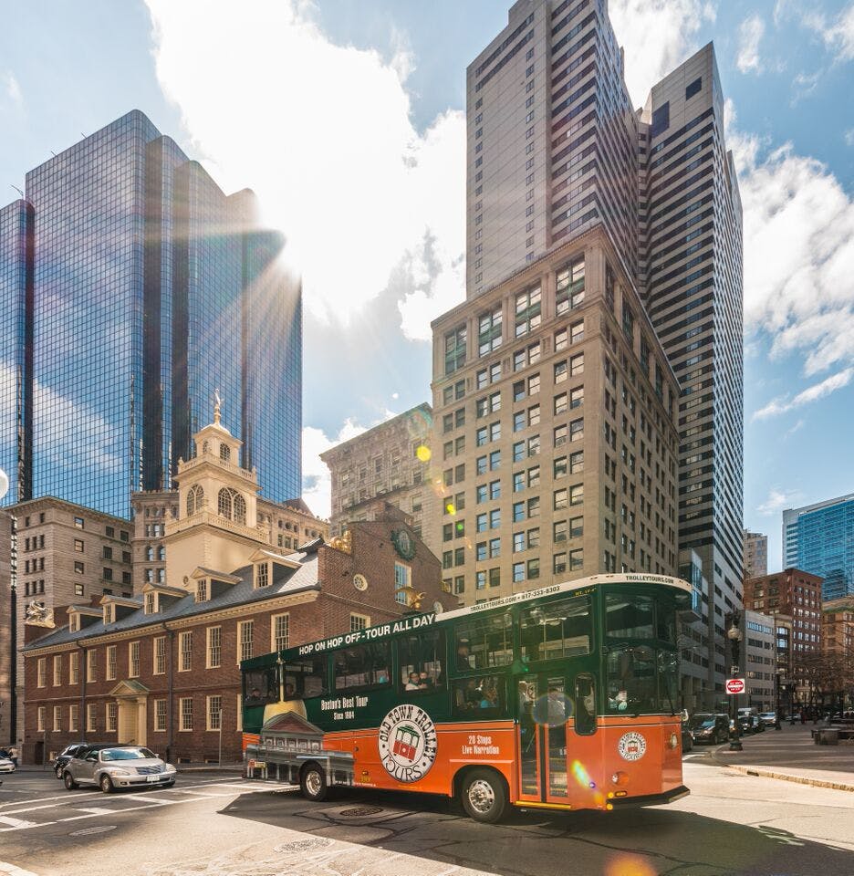 푸른 하늘 아래 현대적인 고층 건물과 역사적인 건물로 둘러싸인 도시 교차로를 오렌지색과 녹색 투어 버스가 운전합니다.