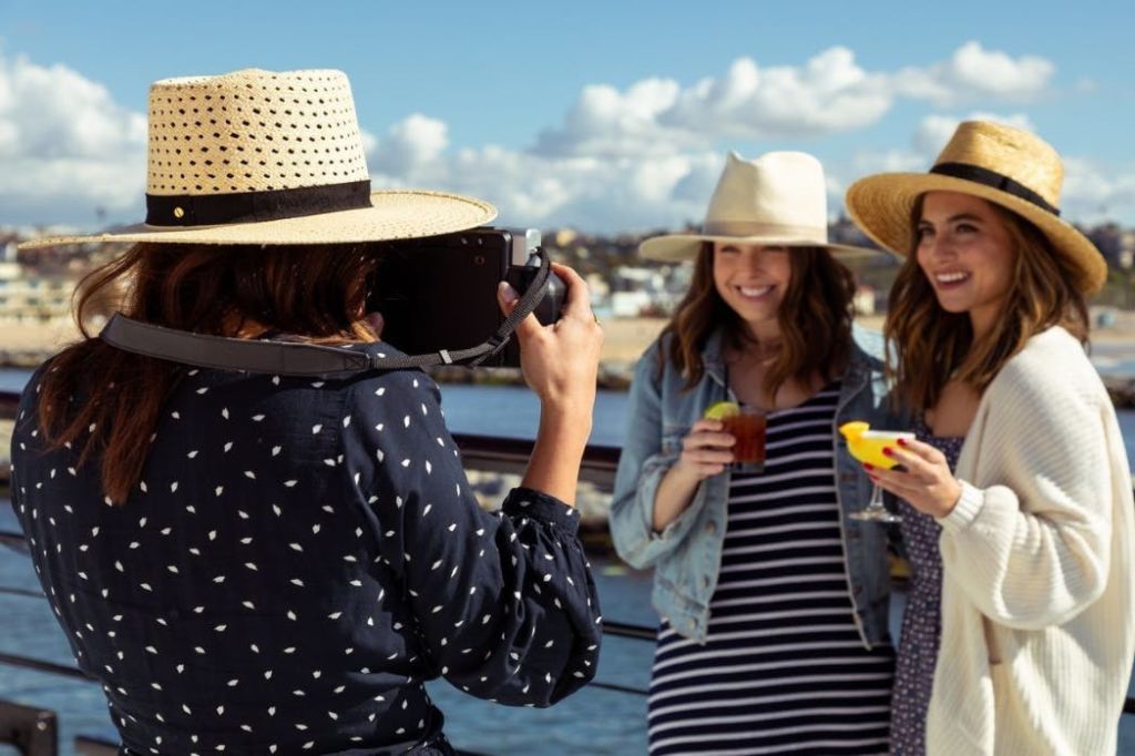 모자를 쓴 두 여성이 카메라를 향해 미소를 짓고 음료를 들고 있으며, 한 여성이 해변을 배경으로 사진을 찍고 있습니다.