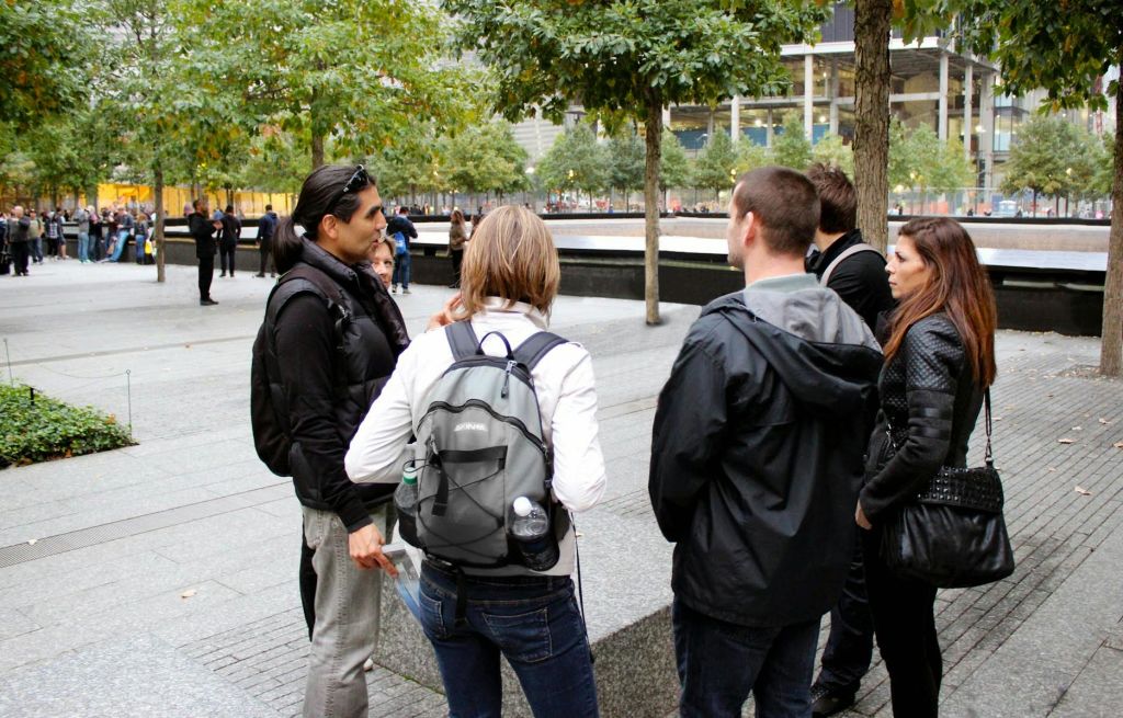 나무와 도시 구조물로 둘러싸인 기념 수영장 근처에 4명이 서 있습니다. 한 사람이 배낭을 가지고 있습니다.