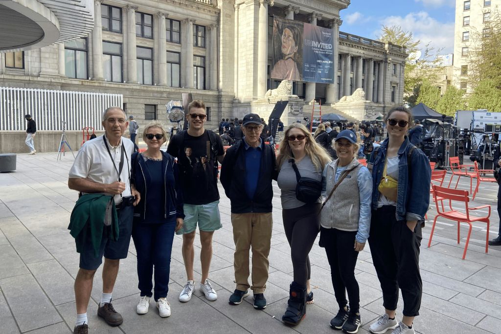 5명이 대형 건물 밖에서 함께 포즈를 취하고 있으며, 배경에는 추가 인원과 촬영 장비가 보입니다.