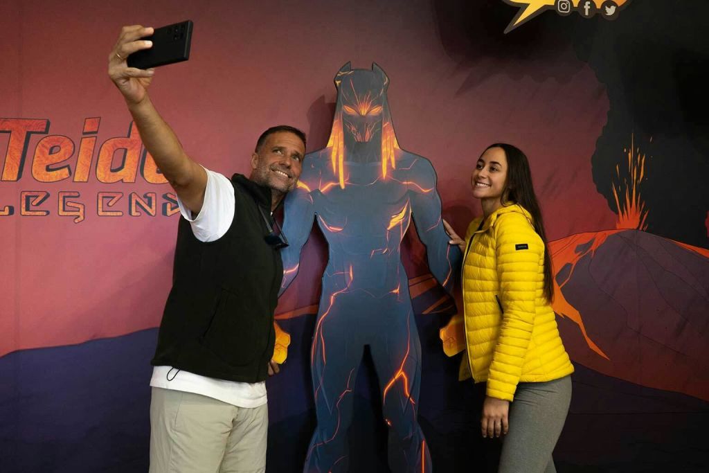 용암을 주제로 한 슈퍼히어로의 컷아웃 옆에서 두 사람이 포즈를 취하고 있습니다. 배경에는 분출하는 화산이 있습니다.