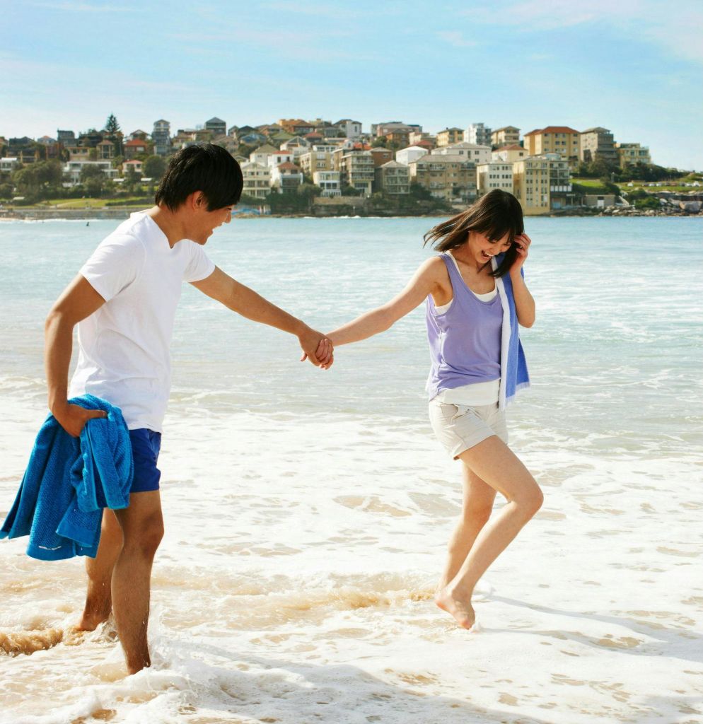 손을 잡고 해변을 거닐고 있는 커플의 발에 물이 튀고 있습니다. 주택이 배경으로 보입니다.