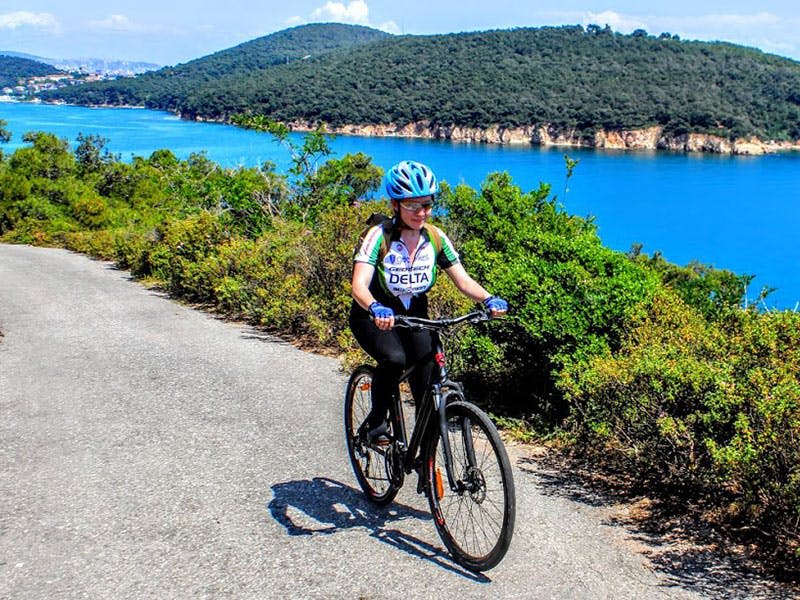 푸르름이 우거진 언덕을 배경으로 활기찬 푸른 물줄기 옆 포장도로에서 자전거를 타는 사람.