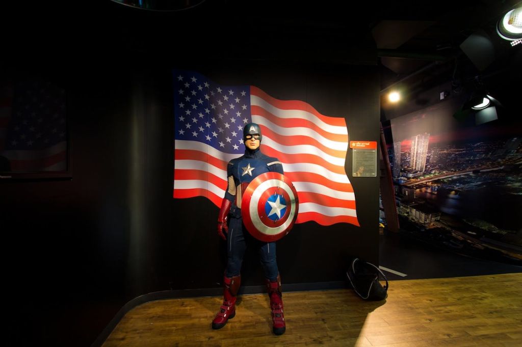 조명이 어두운 방에서 캡틴 아메리카처럼 차려입은 사람이 커다란 미국 국기 배경 앞에 서 있습니다.