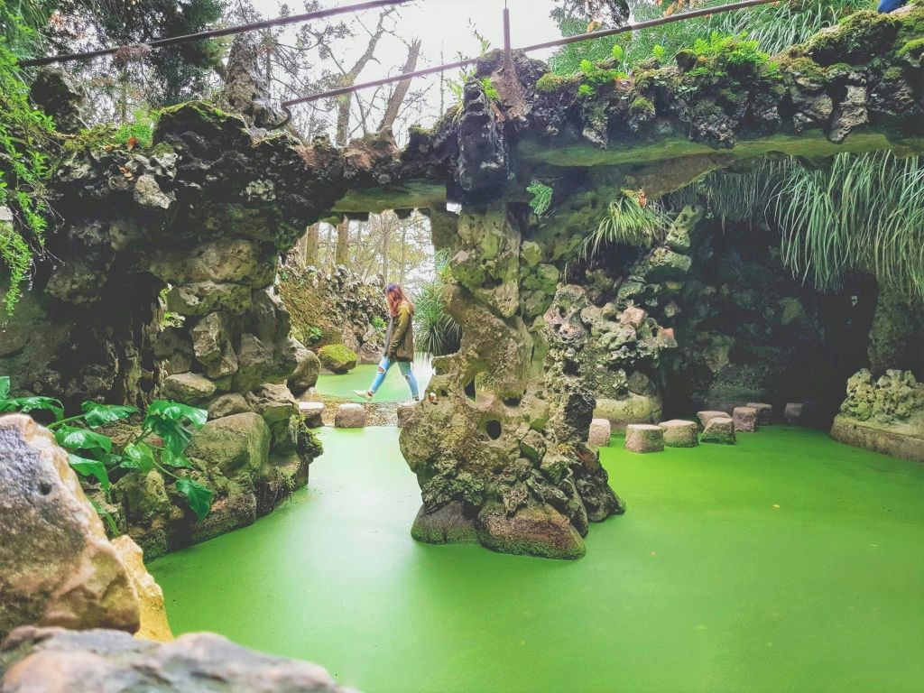 한 사람이 바위처럼 이끼로 뒤덮인 지층과 울창한 녹지로 둘러싸인 푸른 연못 위로 돌계단을 가로질러 걸어가고 있습니다.