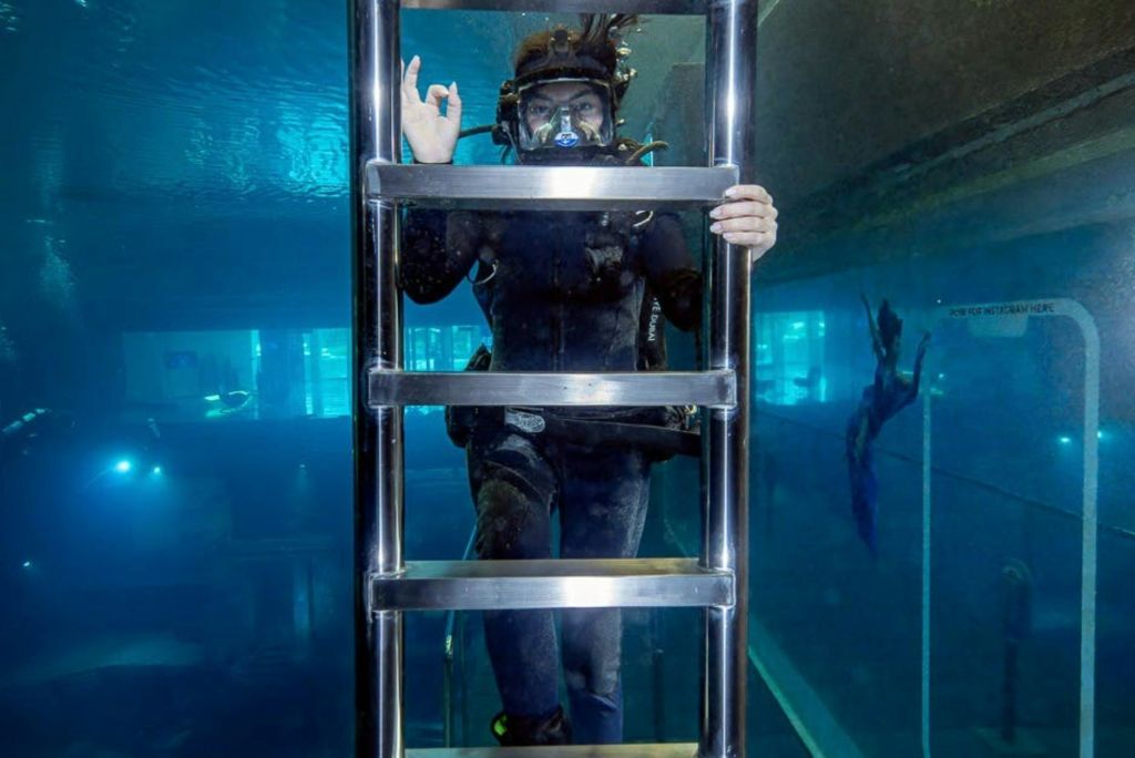 스쿠버 장비를 착용하고 커다란 투명한 물 탱크에서 금속 사다리를 잡고 무난한 제스처를 취하는 수중 사람.