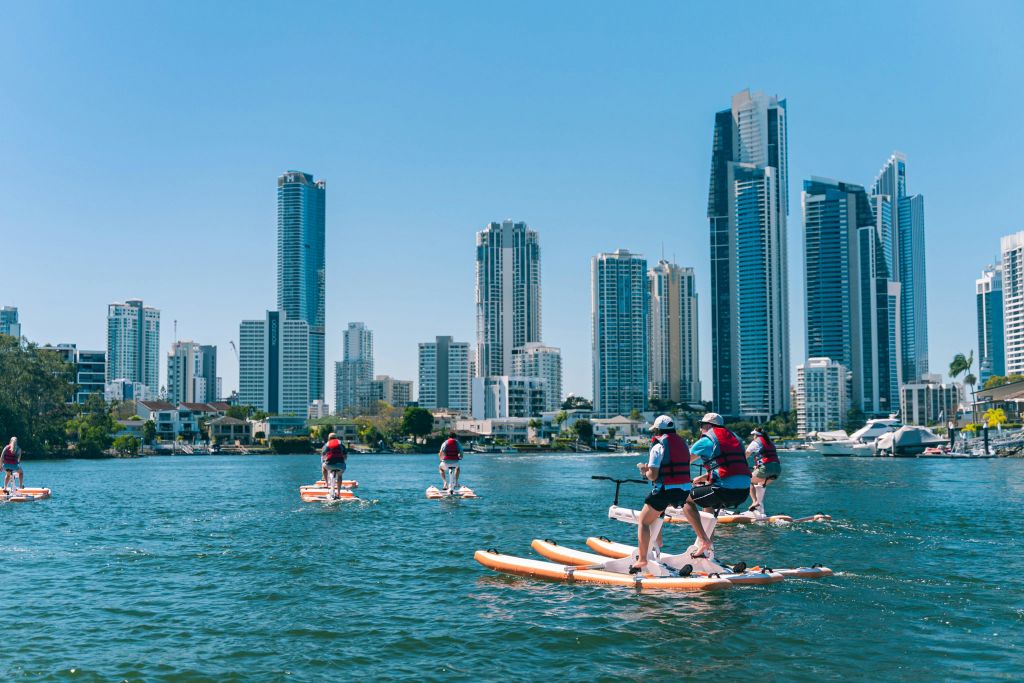 화창한 날, 높은 빌딩을 배경으로 도시의 스카이라인이 펼쳐지는 수로에서 패들보딩을 즐기는 사람들.