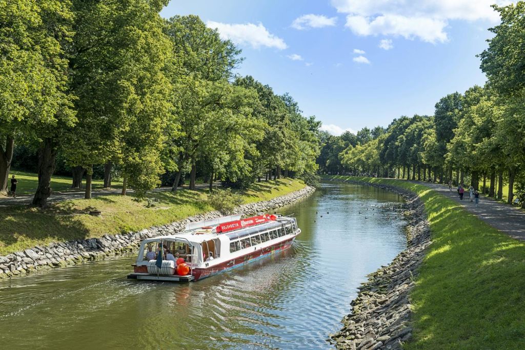 구름이 흩날리는 밝고 푸른 하늘 아래 나무가 늘어선 운하를 따라 관광 보트가 이동합니다.