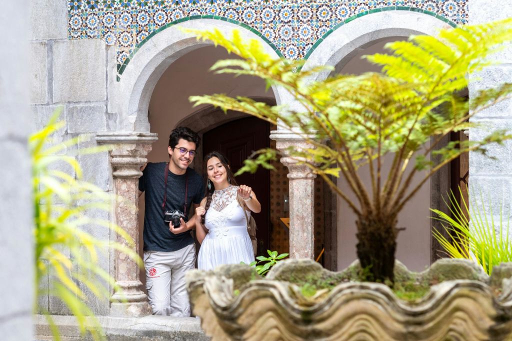 화분과 타일 패턴을 배경으로 장식된 석조 아치길을 배경으로 두 사람이 미소를 짓고 서 있습니다.