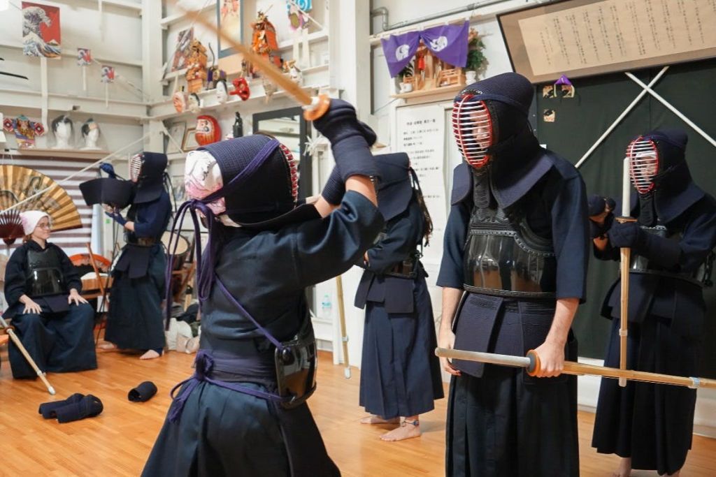 검도 장비를 착용한 두 사람이 도장에서 대나무 검을 들고 연습을 하고 있으며, 백그라운드에는 실무자가 추가로 배치되어 있습니다.