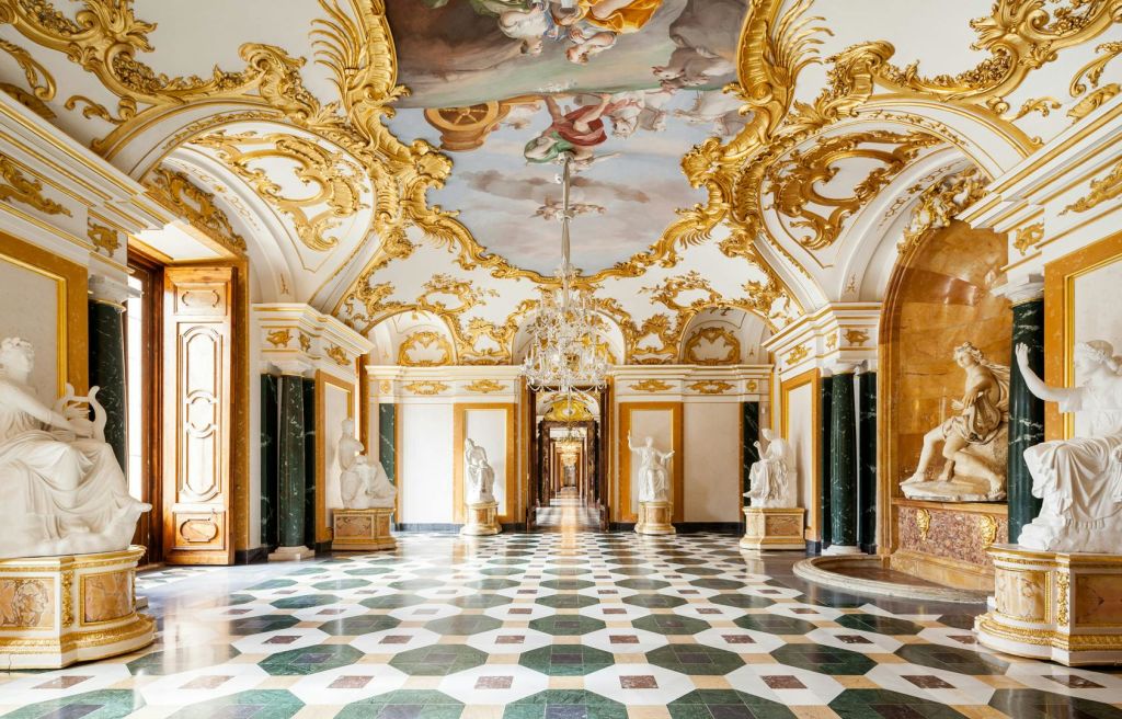 정교한 천장 프레스코화, 골드 디테일, 대리석 조각상, 샹들리에가 있는 화려한 궁전 복도. 기하학적 타일 바닥.