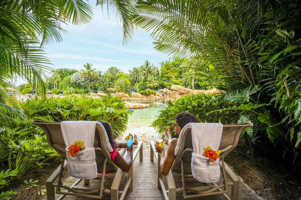 두 사람은 열대 식물로 둘러싸인 음료를 마시며 의자에 앉아 바위와 물이 있는 울창하고 푸른 전망을 감상합니다.