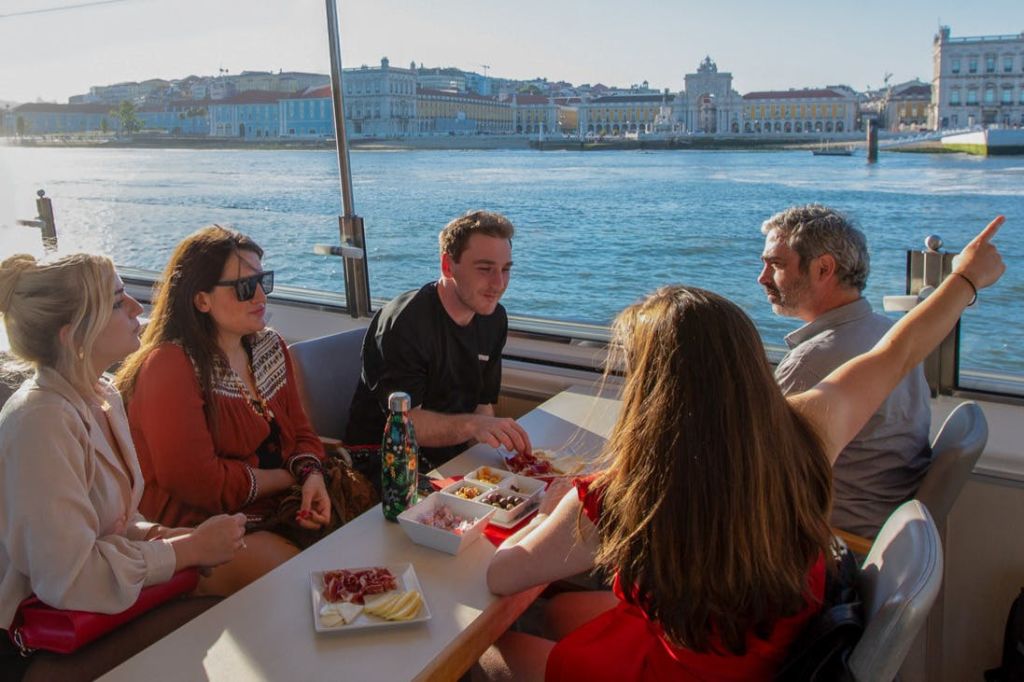 보트 위의 테이블에 앉아 음식을 즐기는 네 명의 사람들, 창문 너머로 도시 풍경과 바닷가가 보입니다.