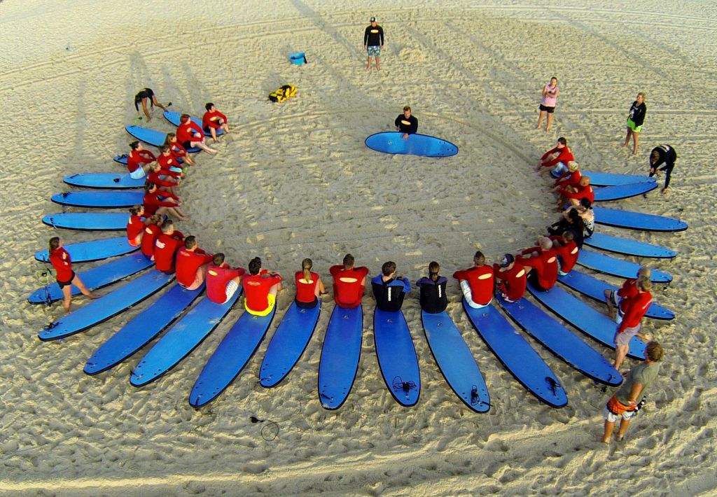 빨간 셔츠를 입은 한 무리의 사람들이 파란색 서핑보드 위에 앉아 모래사장에서 원을 그리며 원 바깥에 세 명의 관찰자가 서 있습니다.