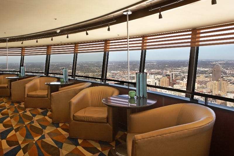 가죽 의자, 작은 테이블, 커다란 창문이 있는 라운지로 탁 트인 도시 전망을 감상할 수 있습니다. 바닥에는 기하학적 패턴의 카펫이 깔려 있습니다.