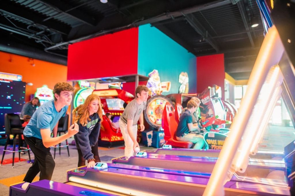 다양한 게임기를 배경으로 화려한 아케이드에서 스키볼을 즐기는 세 사람.