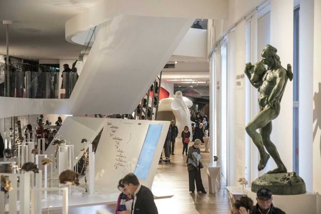 한 남자의 커다란 녹색 청동 동상, 유리 진열장, 돌아다니는 방문객 등 조각품이 있는 박물관 내부.