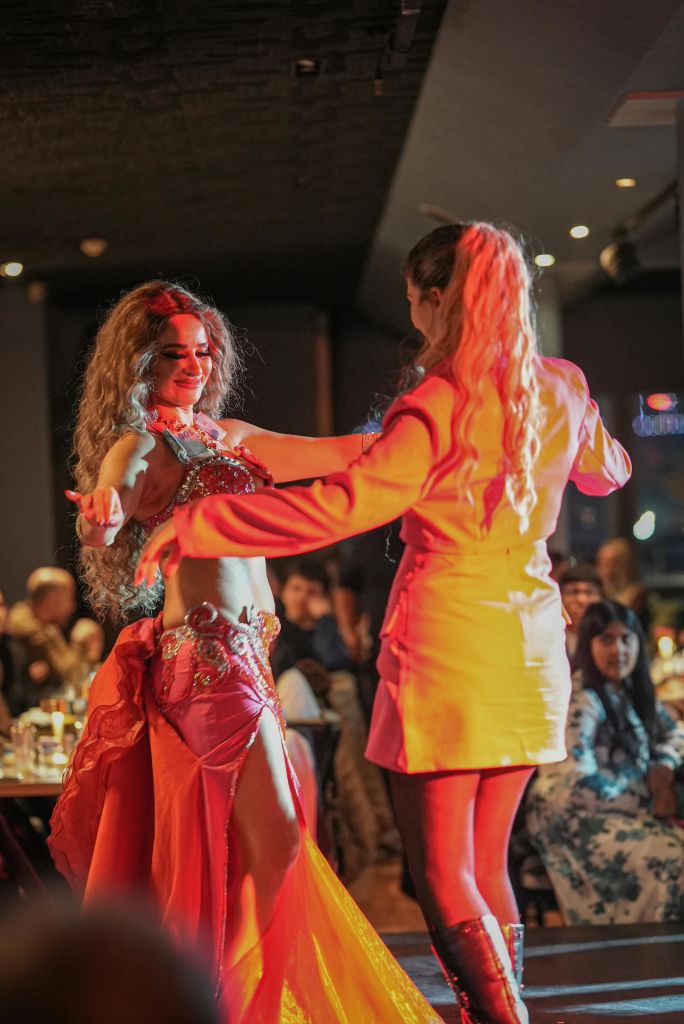 빨간색 의상을 입은 벨리 댄서가 붐비는 조명이 희미한 레스토랑에서 관객과 함께 공연합니다.