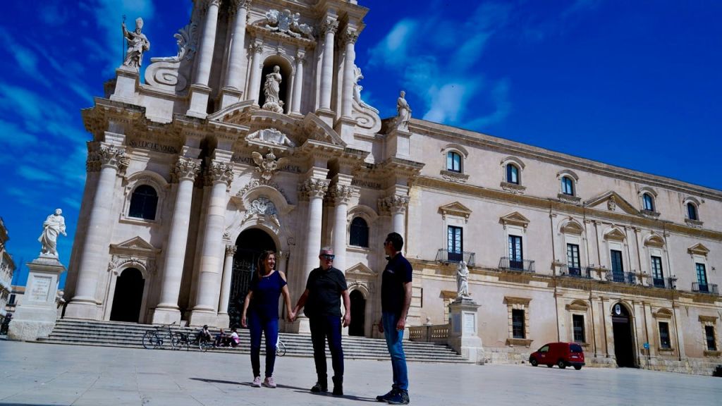 기둥과 조각상이 있는 유서 깊은 건물 앞에 세 사람이 손을 잡고 서 있습니다. 하늘이 맑고 푸르다.