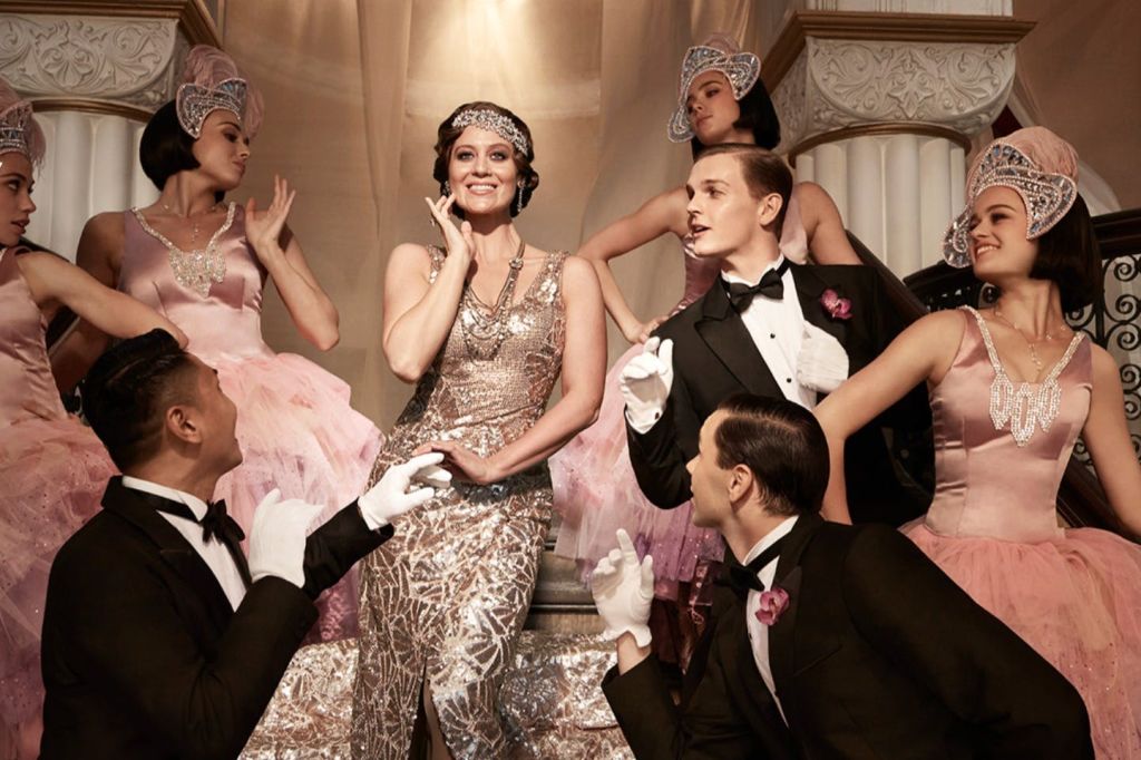 반짝이는 드레스를 입은 매력적인 여성이 웅장한 계단에서 우아하게 차려입은 사람들로 둘러싸여 있습니다.