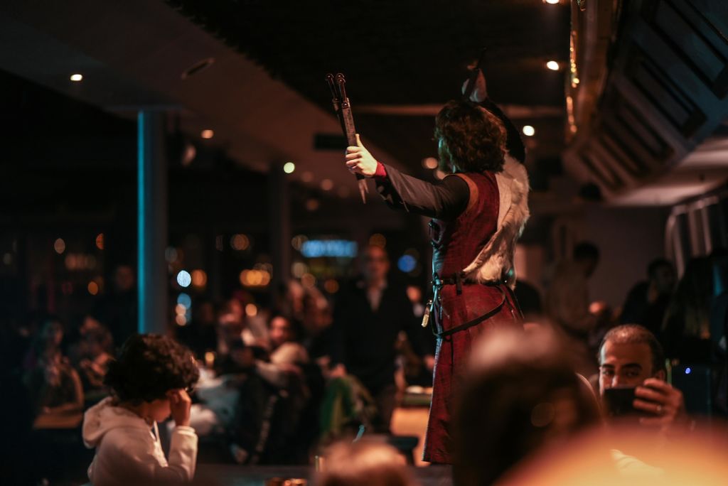 역사적인 복장을 한 사람이 조명이 희미한 방에서 팔을 들어 올리고 관중들이 지켜보고 있습니다.