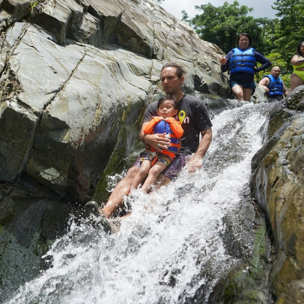 구명조끼를 입은 한 남자와 어린이가 바위 폭포를 따라 미끄러져 내려오고, 위에서 기다리는 다른 사람들이 있습니다. 액티비티를 즐기는 것 같습니다.