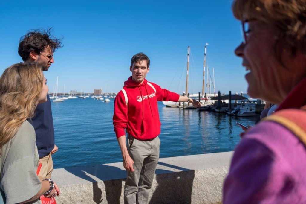 맑고 푸른 하늘 아래 계단식 범선이 있는 바닷가 근처에서 빨간색 후드티를 입은 사람이 다른 사람들과 함께 제스처를 취하고 있습니다.