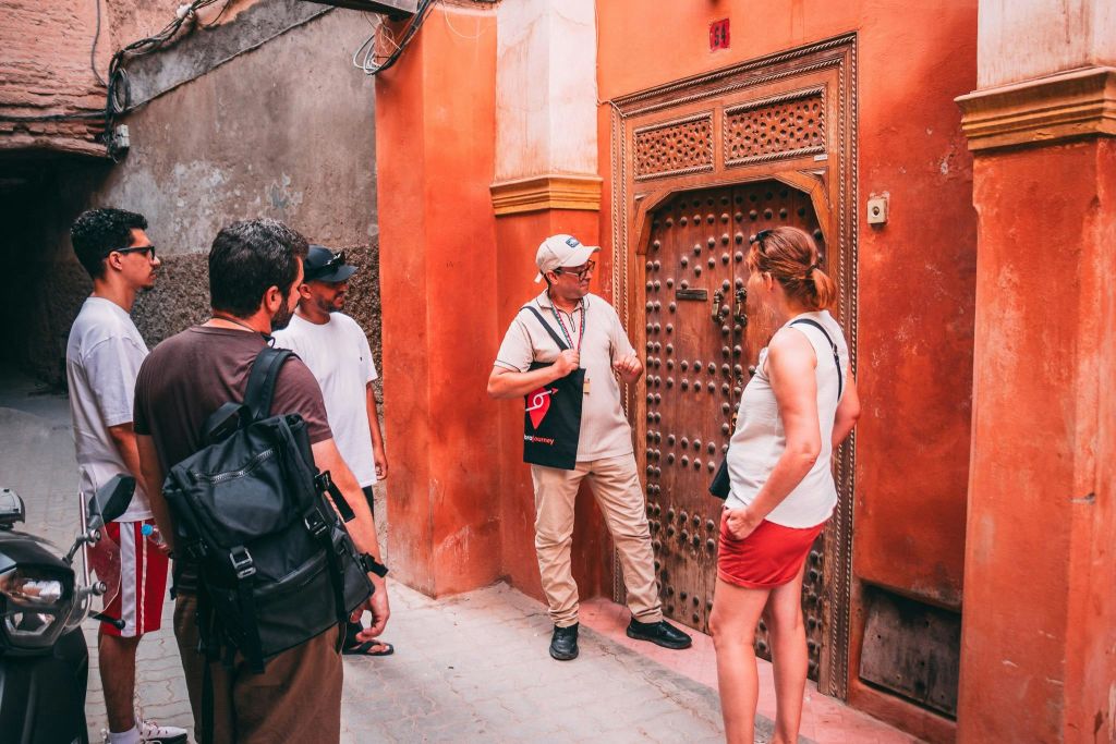 네 명의 남성이 좁은 골목의 화려하고 나무로 된 문 앞에 서서 한 남성이 무언가를 가리키며 설명하고 있습니다.