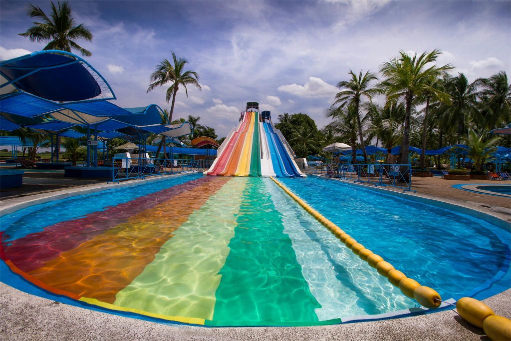 다채로운 다차선 워터 슬라이드가 부분적으로 흐린 하늘 아래 야자수와 푸른 그늘 구조로 둘러싸인 수영장으로 내려갑니다.