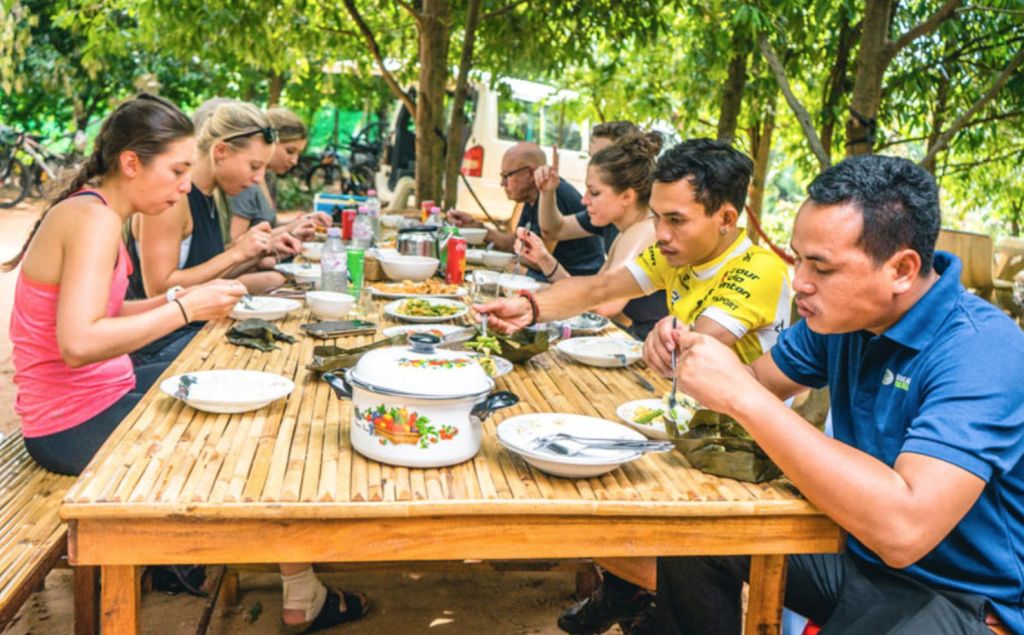4명이 야외의 나무 테이블에 앉아 나무로 둘러싸인 그늘진 공간에서 다양한 요리와 함께 식사를 하고 있습니다.