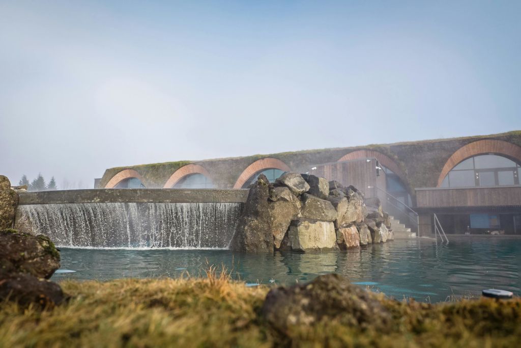 잔디로 덮인 아치형 지붕, 계단식 물, 바위, 전경의 수영장이 있는 현대적인 건물입니다.