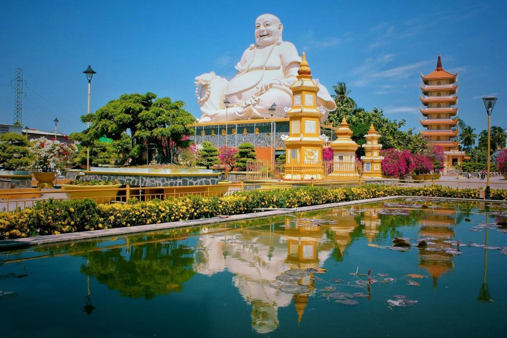 화려한 금빛 구조물과 나무, 푸른 하늘 아래 반사 연못으로 둘러싸인 커다란 흰색 불상.