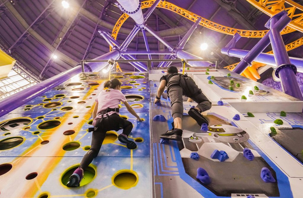 등반 장비를 착용한 두 사람이 높은 천장과 생생한 구조물을 배경으로 다채로운 실내 암벽 등반 벽을 펼칩니다.