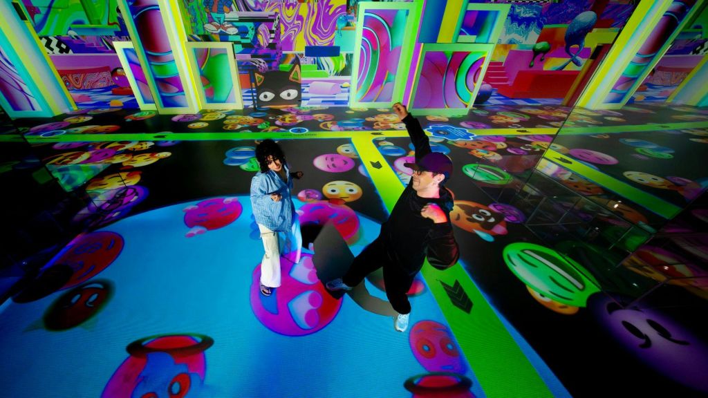 벽과 바닥에 이모티콘과 추상적인 그래픽이 투영된 다채로운 공간에서 두 사람이 상호작용합니다.