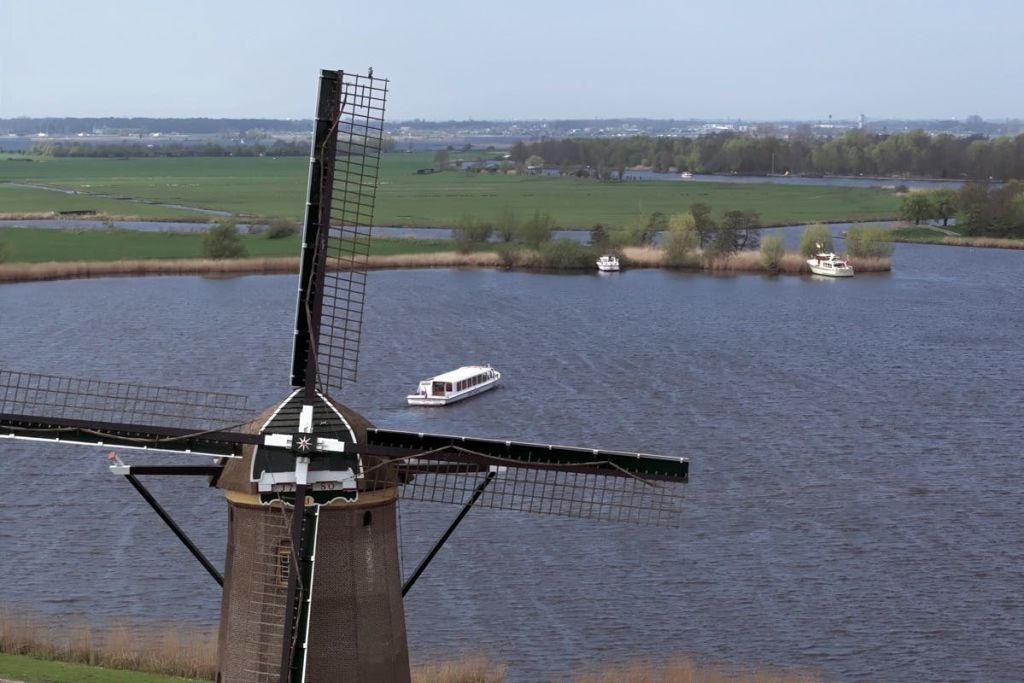 맑은 하늘 아래 물 위에 보트가 있고 푸른 들판을 배경으로 강 근처의 풍차.