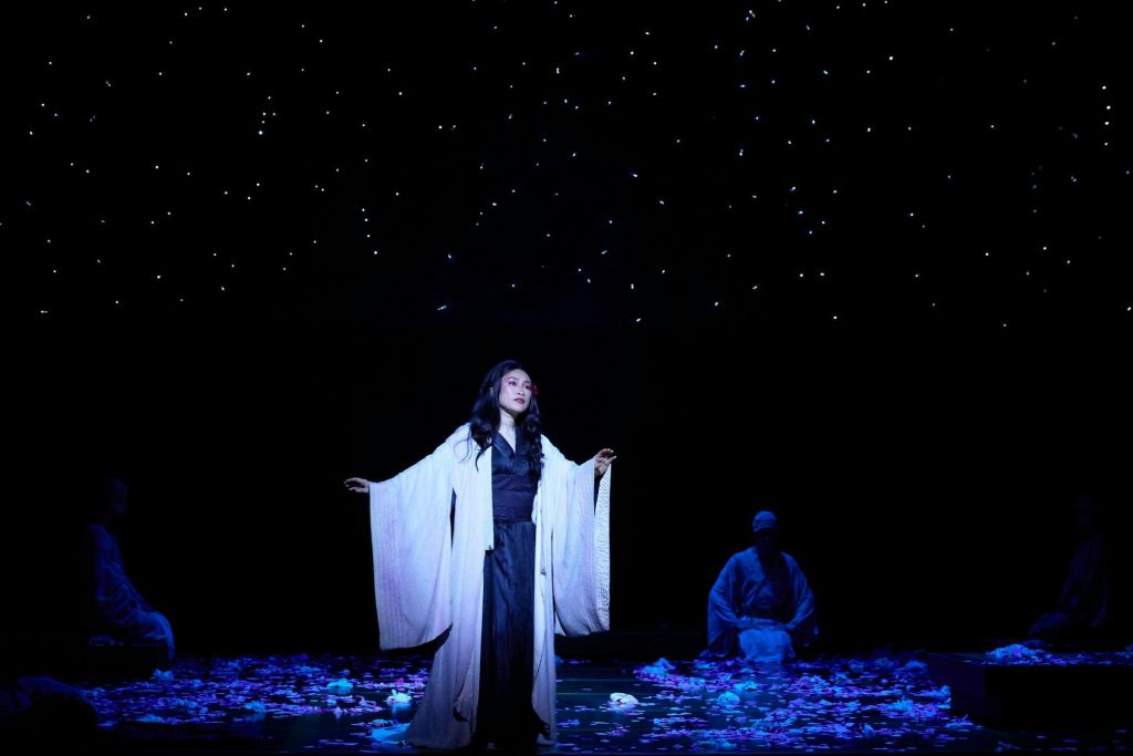 별이 빛나는 무대를 배경으로 흰 가운을 입은 사람이 무대 위에 서 있으며, 배경에는 또 다른 인물이 앉아 있습니다.