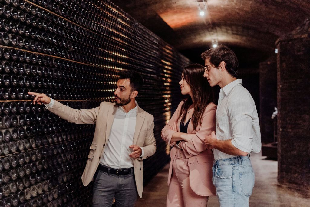 정장 차림의 세 사람이 조명이 어두운 와인 저장고에 서서 선반에 놓인 와인병을 보며 대화를 나누고 있습니다.