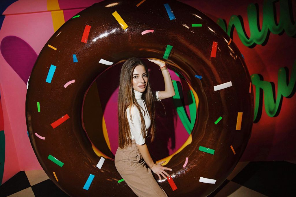 활기찬 분위기에서 다채로운 컬러의 스프링클이 장식된 커다란 원형 초콜릿 도넛 소품을 들고 포즈를 취하고 있는 여성.