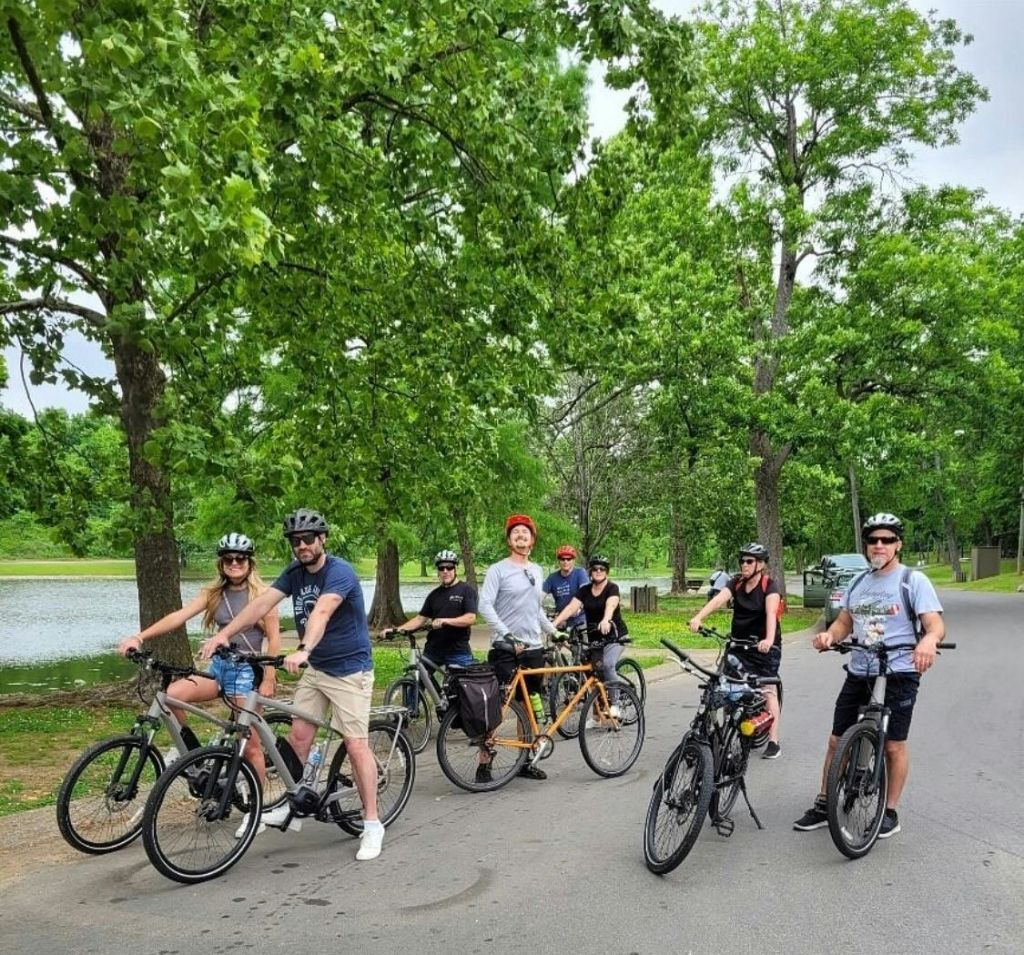 푸른 나무와 작은 호수를 배경으로 한 공원의 포장도로에 자전거를 탄 사람들이 멈춰섰습니다.