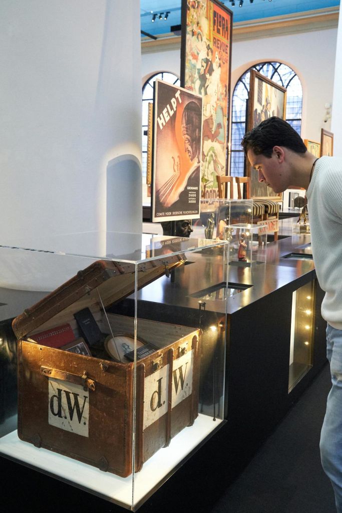 흰색 스웨터를 입은 사람이 박물관의 전시물을 자세히 살펴보고 유리 뒤에 다양한 포스터와 유물을 전시합니다.