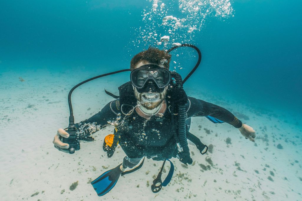 수중에서 잠수복, 마스크, 지느러미를 착용하고 주위에 거품이 솟아오르는 스쿠버 다이버. 아래로 바다 바닥이 보입니다.