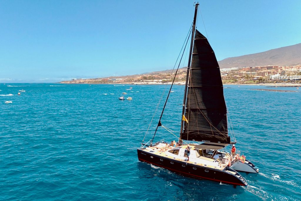 맑고 푸른 바다에서 검은 돛을 단 범선으로, 햇살 가득한 하늘 아래 배경에 해안선이 보입니다.